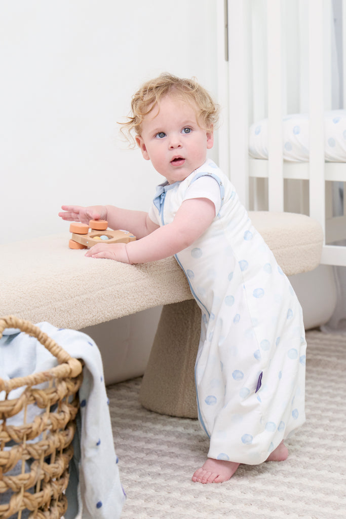 Child in a blue dot clevasleep dreamsuit standing next to a beige stool in a nursery.