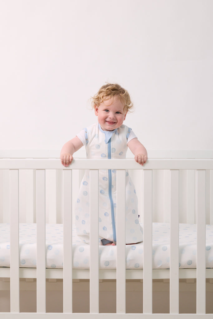 Child standing in a crib wearing a blue dot clevasleep dreamsuit with a white background